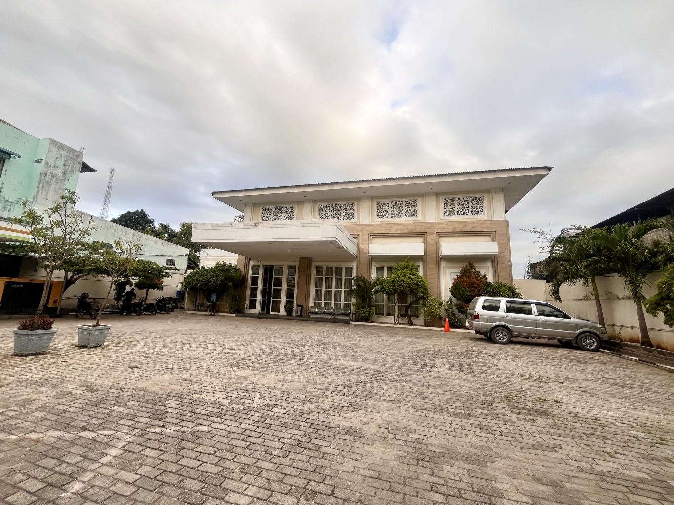 The beige and white exterior of Ella Hotel in Tambolaka features a wide paved entrance, with a silver SUV parked on the right under an overcast sky.