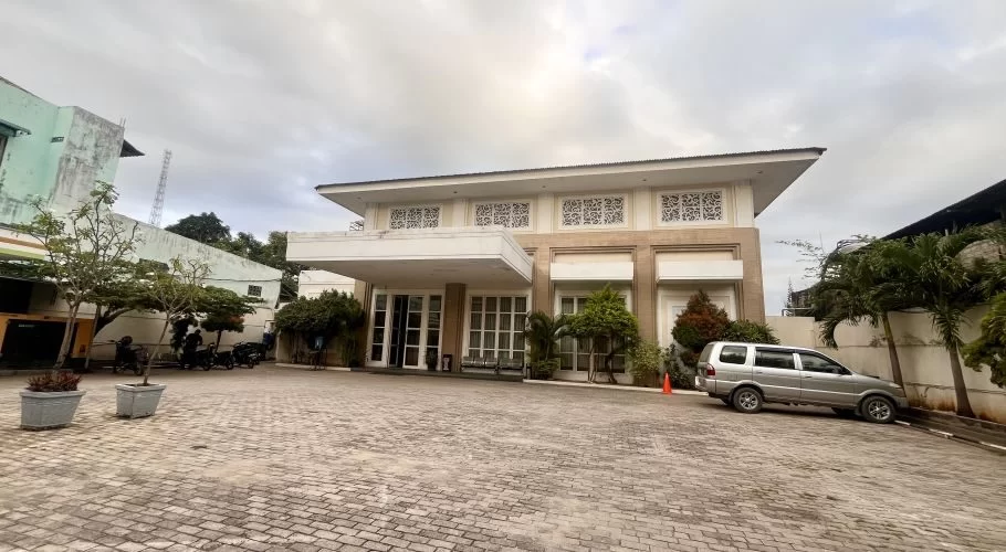 The beige and white exterior of Ella Hotel in Tambolaka features a wide paved entrance, with a silver SUV parked on the right under an overcast sky.