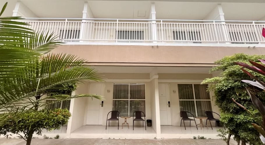 A two-story hotel building with a light brown and white facade, featuring ground-floor rooms with seating and a second-floor balcony, surrounded by green tropical plants.