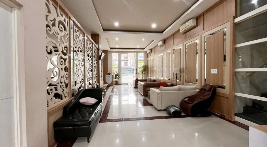 A spacious hotel lobby features white and brown tiled floors, light wood paneled walls, various sofas, a black chaise lounge, and a front desk area leading to glass entrance doors.