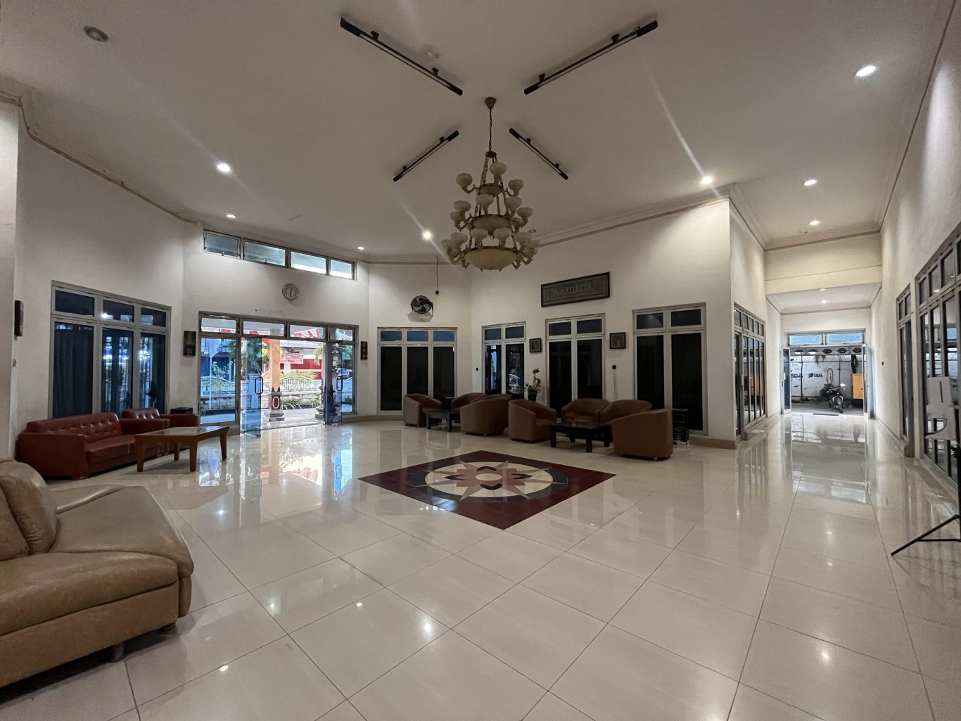 A spacious, brightly lit hotel lobby features white walls, shiny light-colored tiled floors with a central pattern, and multiple brown seating arrangements.