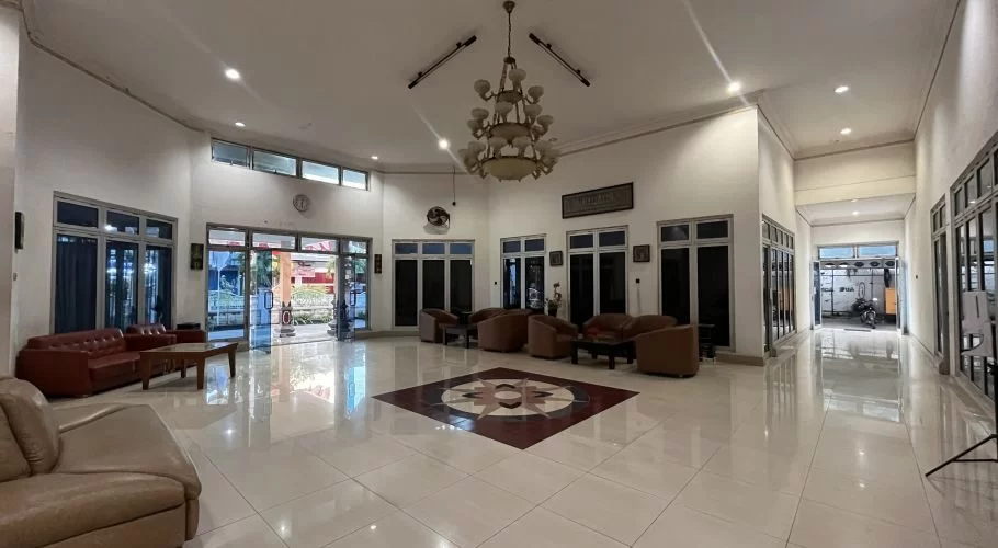 A spacious, brightly lit hotel lobby features white walls, shiny light-colored tiled floors with a central pattern, and multiple brown seating arrangements.