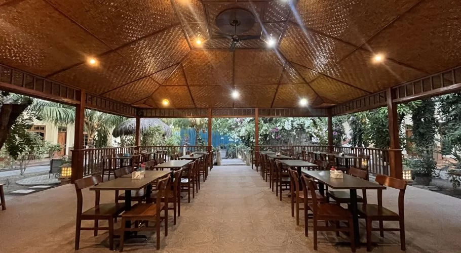 A spacious, open-air restaurant at Elvin Hotel features a high woven ceiling, dark wooden tables, and lush greenery outside.
