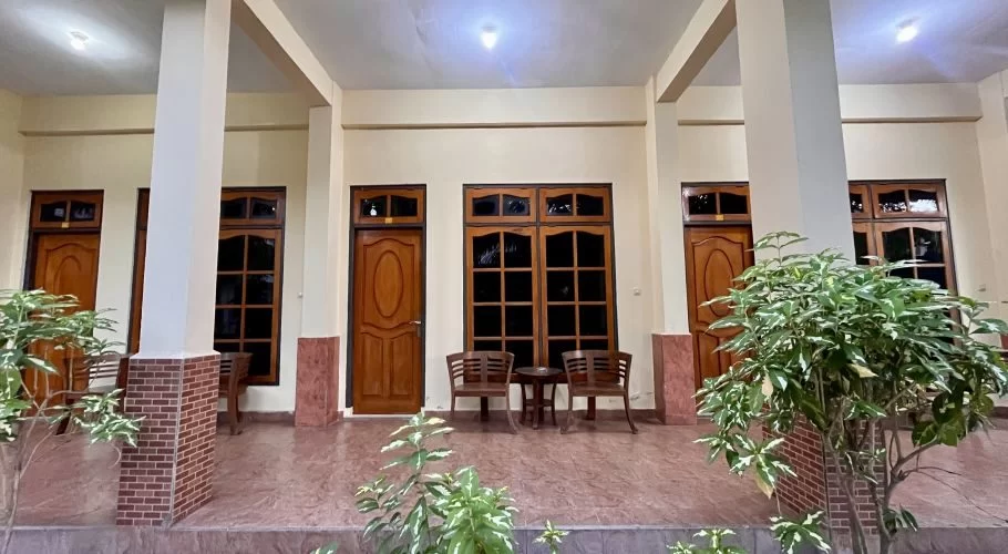 A traditional hotel corridor features beige walls, brown wooden doors and windows, white pillars, and potted green plants, with two chairs and a small table.