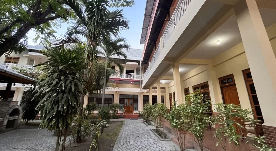 A traditional hotel courtyard features light-colored buildings with balconies, a paved walkway, and lush tropical plants under a clear sky.