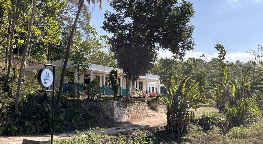 The white and green English Goes to Kampung (EGK) building stands amidst dense tropical foliage under a clear blue sky in rural Sumba.