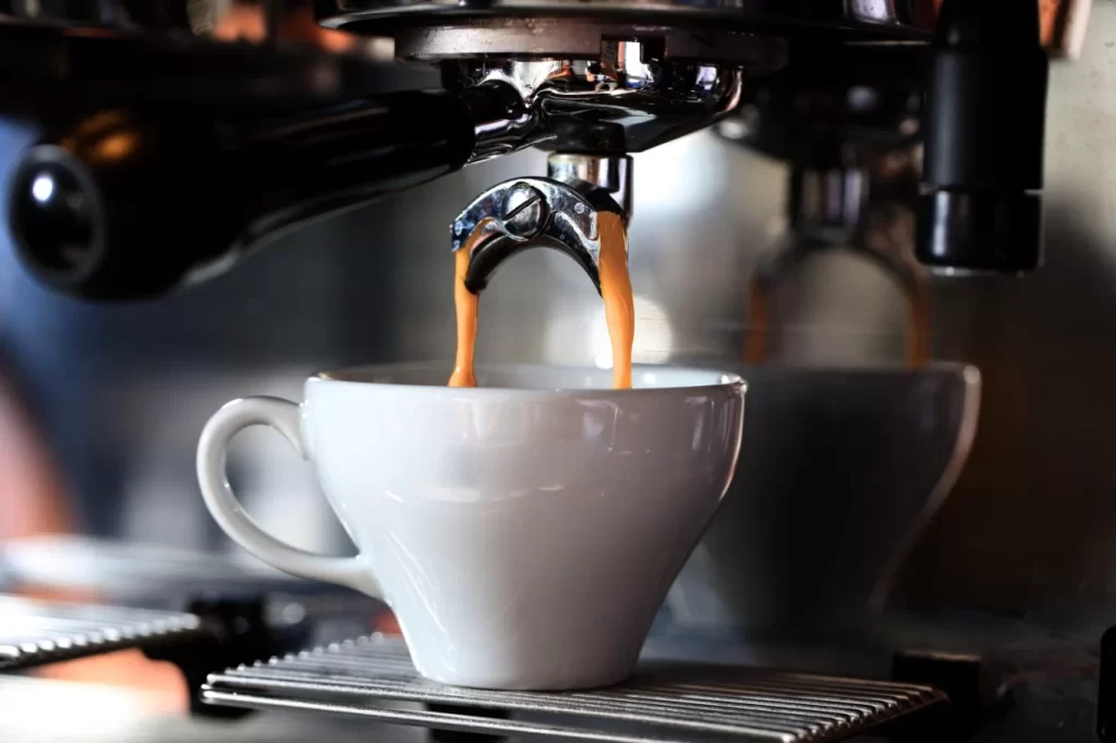 Espresso streams into a white cup from a chrome espresso machine's portafilter, set on a metal grate.