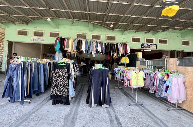 Inside the Gajah Mada thrift store in Waikabubak, numerous racks of colorful second-hand clothing are displayed under a corrugated metal roof and light green walls.