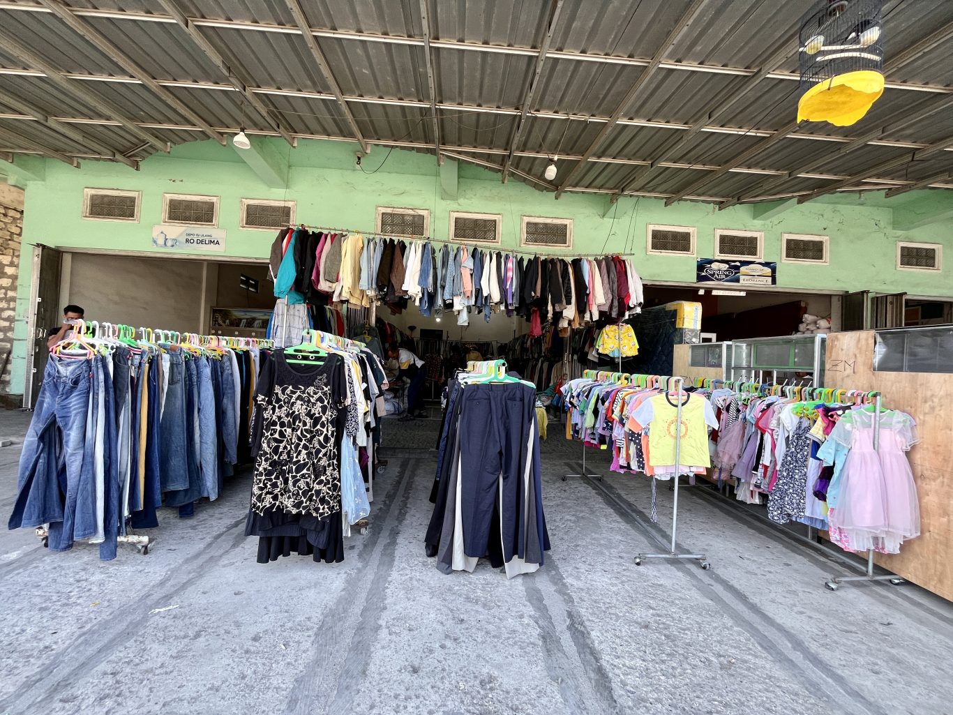 Inside the Gajah Mada thrift store in Waikabubak, numerous racks of colorful second-hand clothing are displayed under a corrugated metal roof and light green walls.
