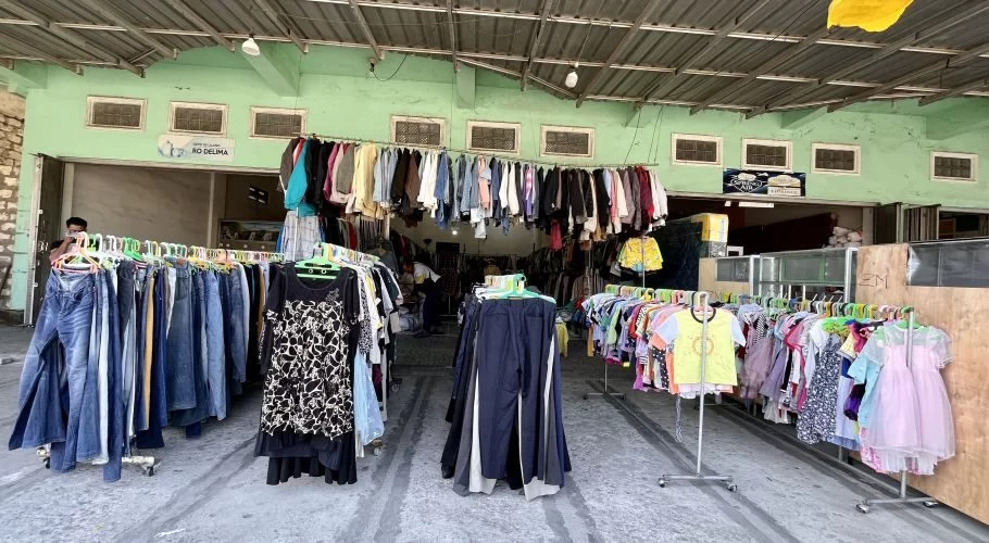 Inside the Gajah Mada thrift store in Waikabubak, numerous racks of colorful second-hand clothing are displayed under a corrugated metal roof and light green walls.