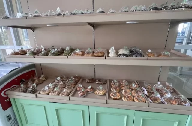 Bakery shelves display numerous individually packaged pastries and donuts on trays, next to a red and white ice cream freezer, within a shop featuring white and green accents.
