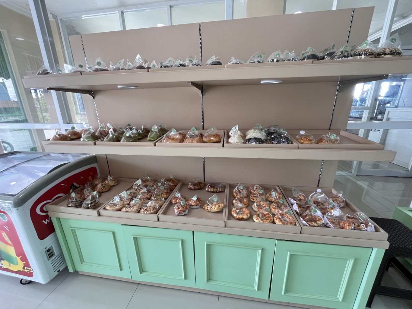 Bakery shelves display numerous individually packaged pastries and donuts on trays, next to a red and white ice cream freezer, within a shop featuring white and green accents.