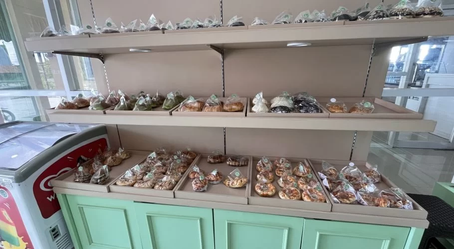 Bakery shelves display numerous individually packaged pastries and donuts on trays, next to a red and white ice cream freezer, within a shop featuring white and green accents.