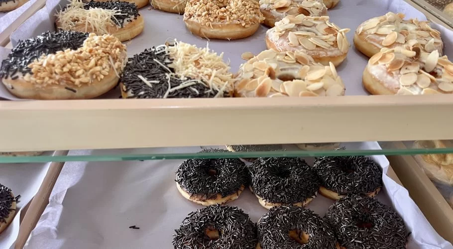 Visible are two shelves of a bakery display case filled with various donuts topped with chocolate sprinkles, peanuts, shredded cheese, and sliced almonds.