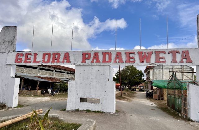 A white concrete archway with "GELORA PADAEWETA" in red letters stands under a partly cloudy sky, marking the entrance to a rural venue with buildings and a dirt road.