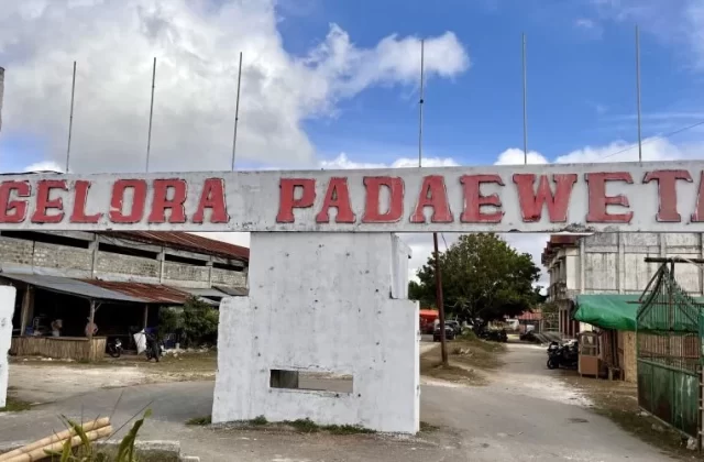 A white concrete archway with "GELORA PADAEWETA" in red letters stands under a partly cloudy sky, marking the entrance to a rural venue with buildings and a dirt road.