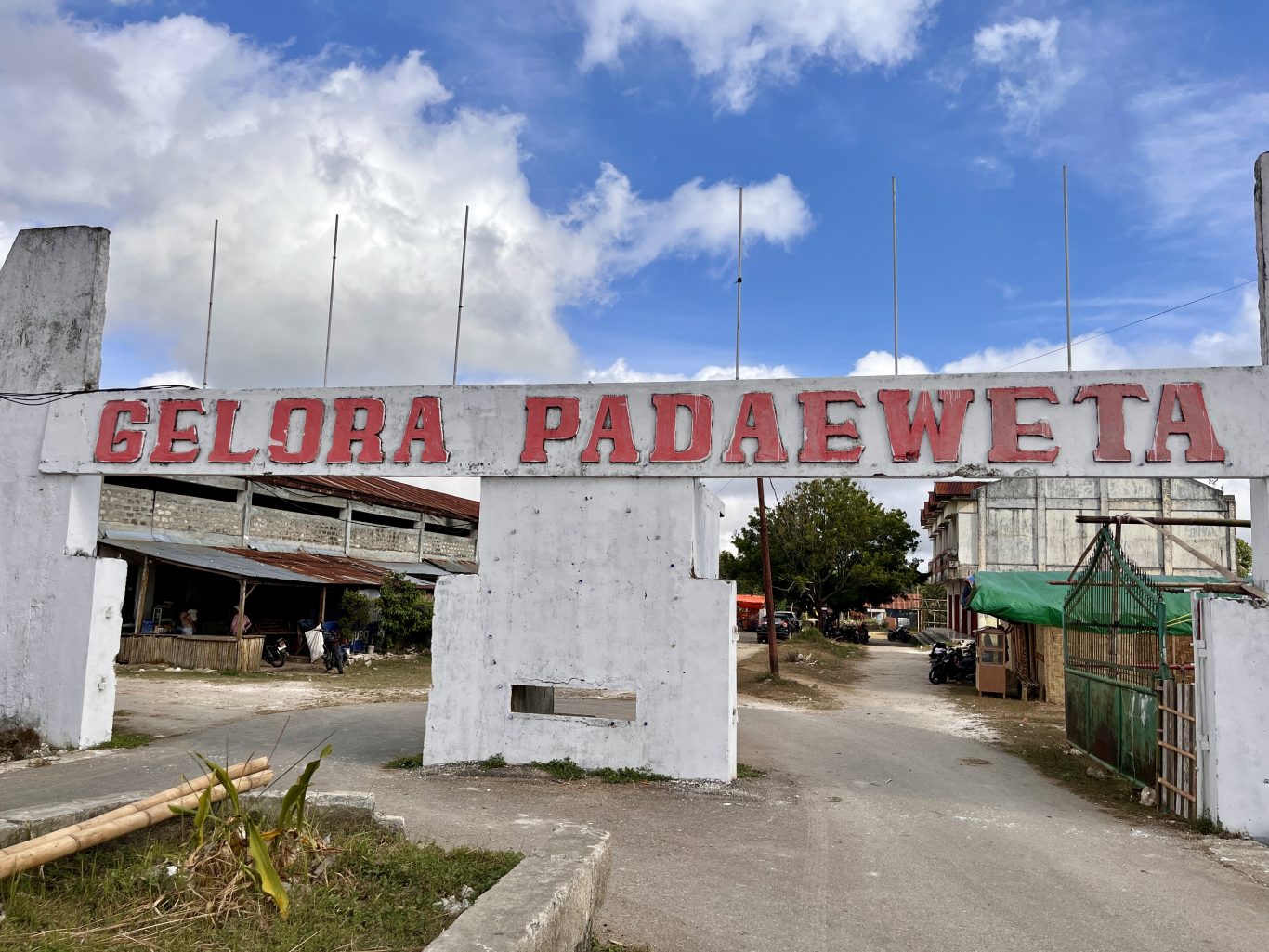 A white concrete archway with "GELORA PADAEWETA" in red letters stands under a partly cloudy sky, marking the entrance to a rural venue with buildings and a dirt road.