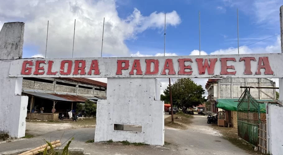A white concrete archway with "GELORA PADAEWETA" in red letters stands under a partly cloudy sky, marking the entrance to a rural venue with buildings and a dirt road.