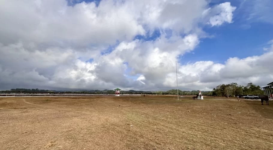 A wide view of the Gelora Pada Eweta horse racing venue in Sumba, featuring a dry brown track, a small red-roofed watchtower, and a partly cloudy blue sky.