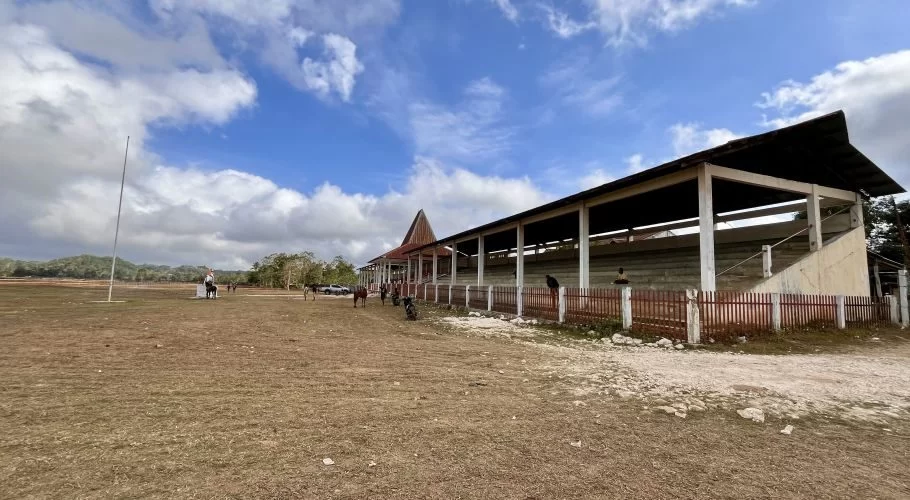 A wide view of the Gelora Pada Eweta horse race track in Sumba, featuring a long grandstand, a dry brown field, and distant green hills under a blue, cloudy sky.