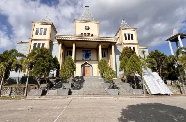 A grand cream and yellow church with a cross-topped spire stands under a partly cloudy sky, flanked by palm trees and featuring a large open concrete Bible monument on the right.