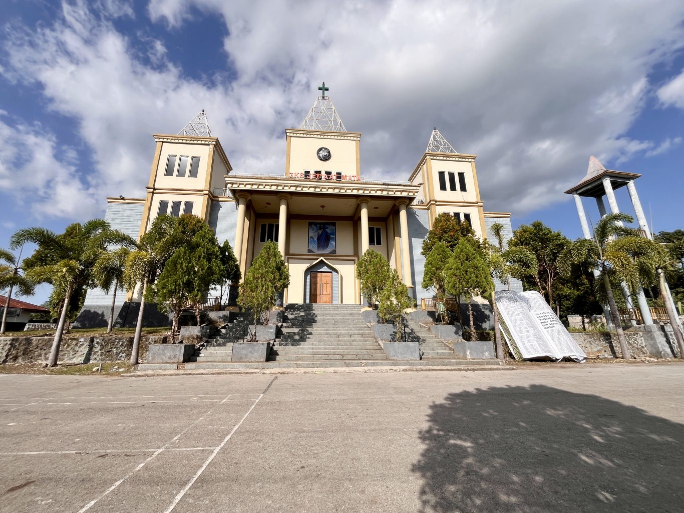 A grand cream and yellow church with a cross-topped spire stands under a partly cloudy sky, flanked by palm trees and featuring a large open concrete Bible monument on the right.