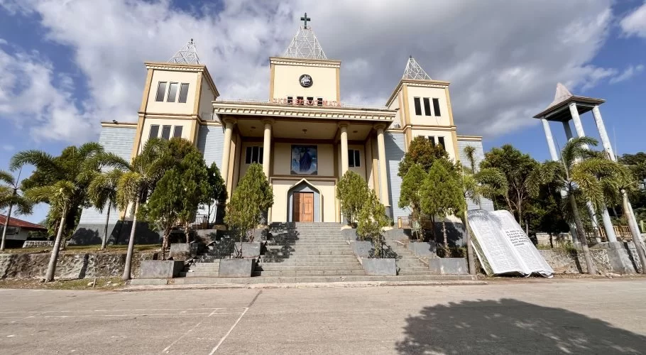 A grand cream and yellow church with a cross-topped spire stands under a partly cloudy sky, flanked by palm trees and featuring a large open concrete Bible monument on the right.