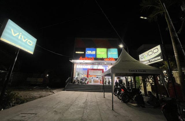 The Global Teknik electronics store in Waikabubak glows brightly at night, displaying brand signs like Vivo and Samsung, with motorcycles parked near an outdoor tent.