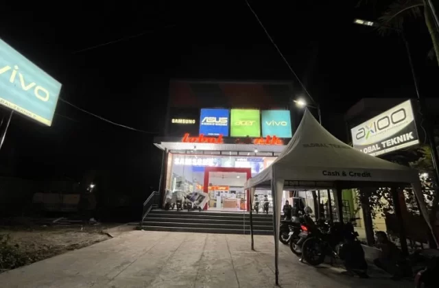 The Global Teknik electronics store in Waikabubak glows brightly at night, displaying brand signs like Vivo and Samsung, with motorcycles parked near an outdoor tent.