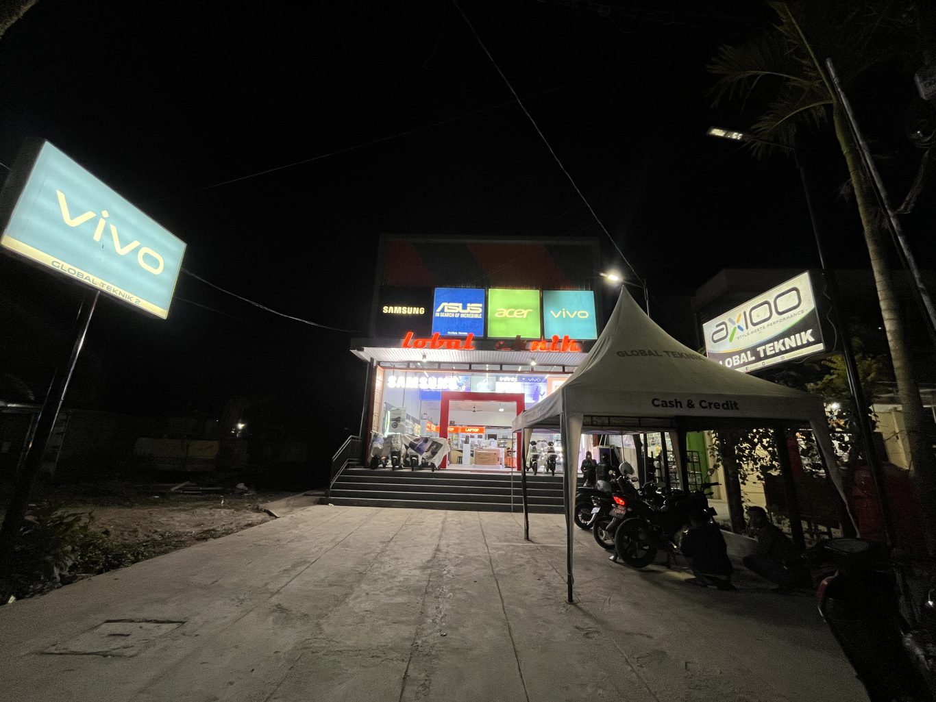 The Global Teknik electronics store in Waikabubak glows brightly at night, displaying brand signs like Vivo and Samsung, with motorcycles parked near an outdoor tent.