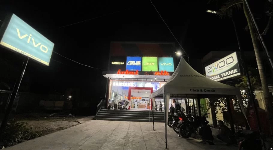 The Global Teknik electronics store in Waikabubak glows brightly at night, displaying brand signs like Vivo and Samsung, with motorcycles parked near an outdoor tent.