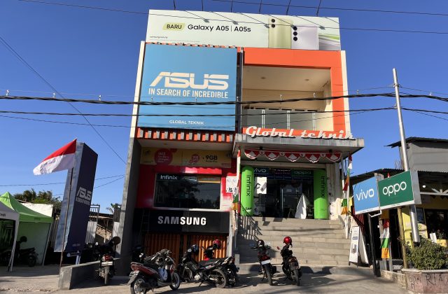 The two-story Global Teknik electronics store in Waingapu, Sumba, features prominent brand signs, an Indonesian flag, and parked motorcycles under a clear blue sky.