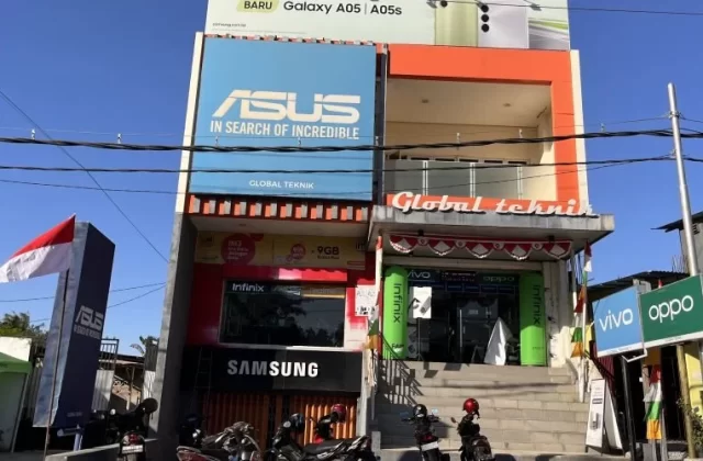 The two-story Global Teknik electronics store in Waingapu, Sumba, features prominent brand signs, an Indonesian flag, and parked motorcycles under a clear blue sky.