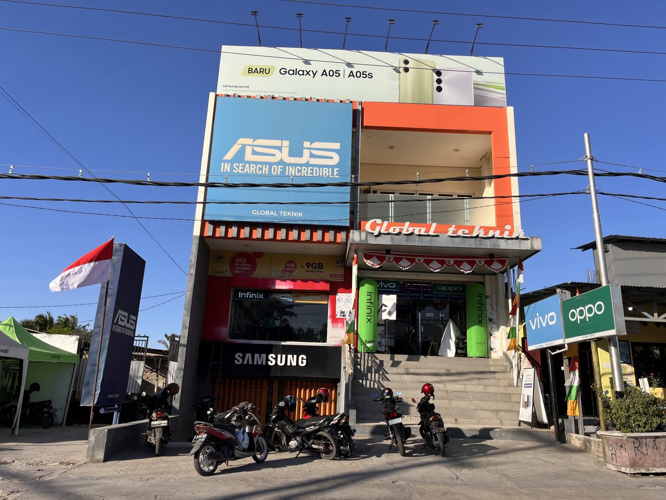 The two-story Global Teknik electronics store in Waingapu, Sumba, features prominent brand signs, an Indonesian flag, and parked motorcycles under a clear blue sky.