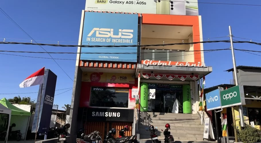 The two-story Global Teknik electronics store in Waingapu, Sumba, features prominent brand signs, an Indonesian flag, and parked motorcycles under a clear blue sky.