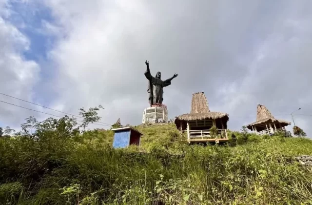 A large dark Jesus statue with outstretched arms stands on a white base atop a green, grassy hill under a cloudy sky, flanked by two thatched gazebos.
