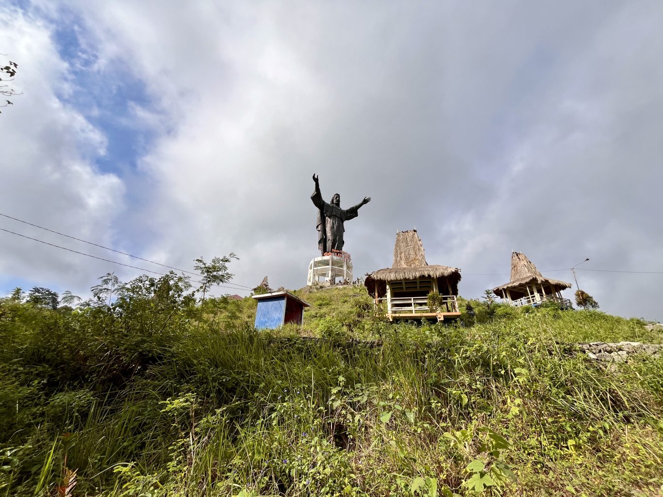 A large dark Jesus statue with outstretched arms stands on a white base atop a green, grassy hill under a cloudy sky, flanked by two thatched gazebos.