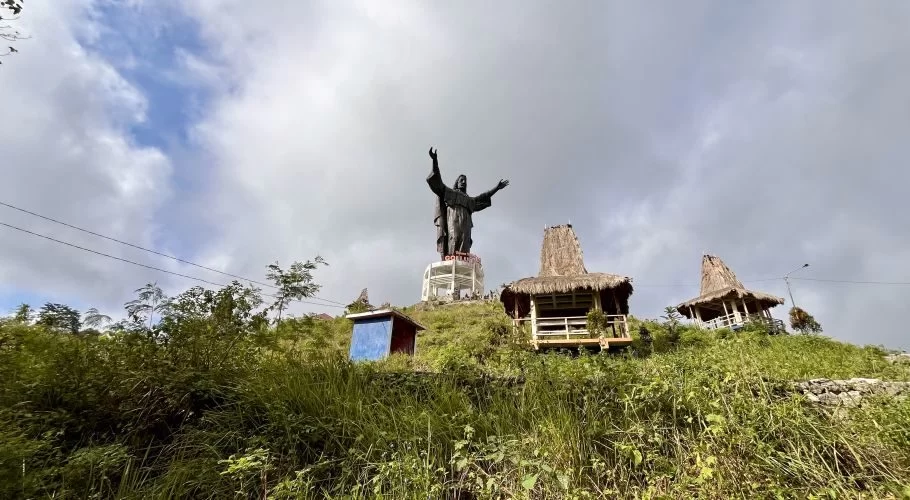 A large dark Jesus statue with outstretched arms stands on a white base atop a green, grassy hill under a cloudy sky, flanked by two thatched gazebos.