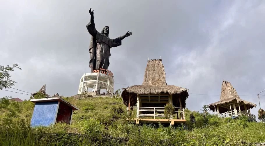 A large dark Jesus statue with outstretched arms stands on a white base atop a grassy hill, flanked by traditional thatched-roof gazebos under a cloudy sky.