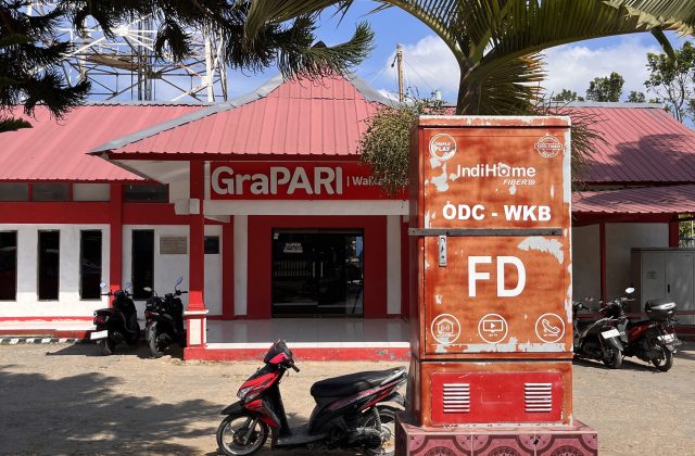 A red and white GraPARI Telkomsel building in Waikabubak, Sumba, with a large IndiHome fiber box and several motorbikes parked in front under a blue sky.