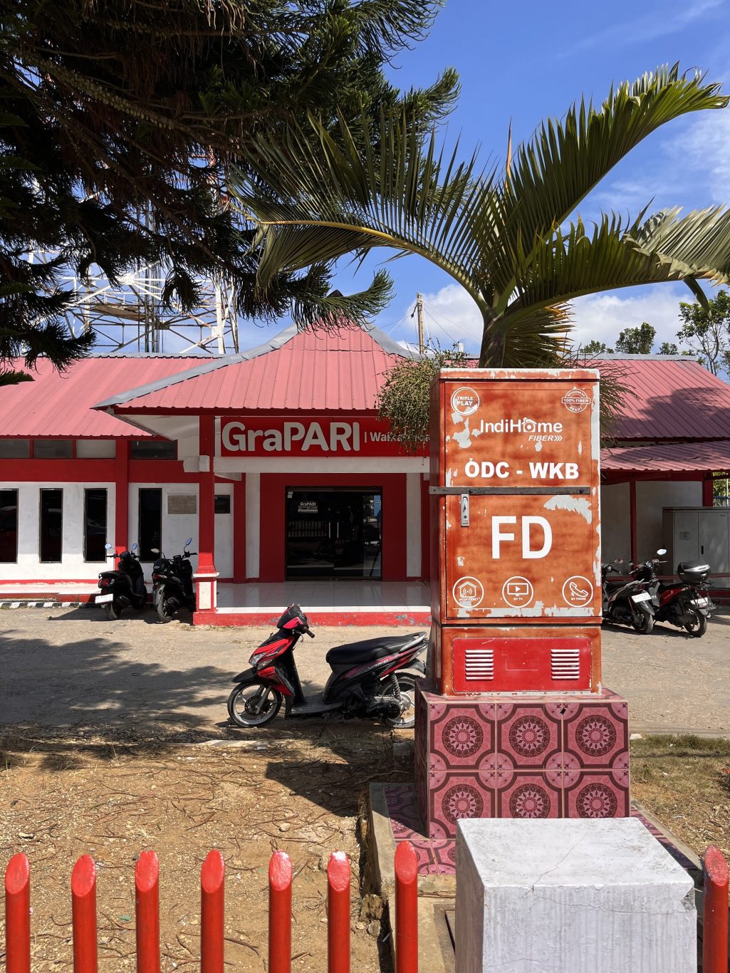 A red and white GraPARI Telkomsel building in Waikabubak, Sumba, with a large IndiHome fiber box and several motorbikes parked in front under a blue sky.