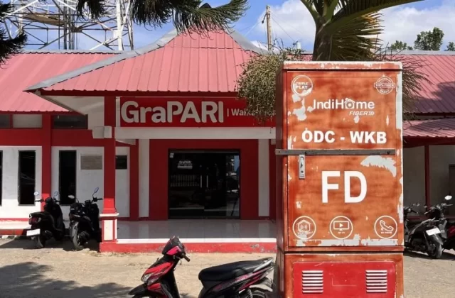 A red and white GraPARI Telkomsel building in Waikabubak, Sumba, with a large IndiHome fiber box and several motorbikes parked in front under a blue sky.