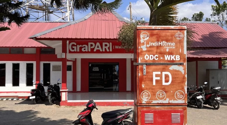 A red and white GraPARI Telkomsel building in Waikabubak, Sumba, with a large IndiHome fiber box and several motorbikes parked in front under a blue sky.