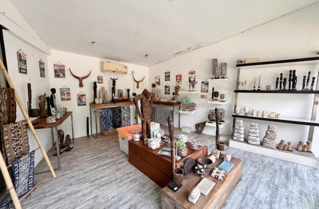 A bright, white-walled art shop interior in Sumba displays numerous traditional wooden carvings, woven textiles, and decorative artifacts on shelves and tables.