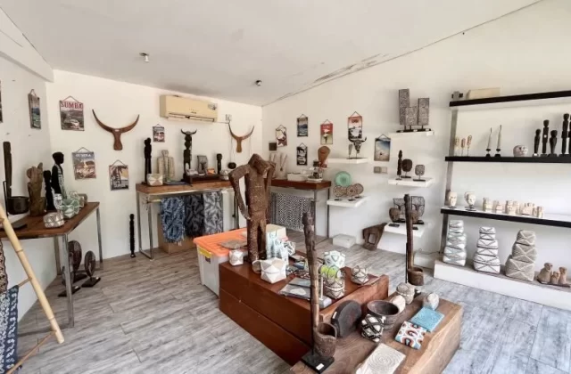 A bright, white-walled art shop interior in Sumba displays numerous traditional wooden carvings, woven textiles, and decorative artifacts on shelves and tables.