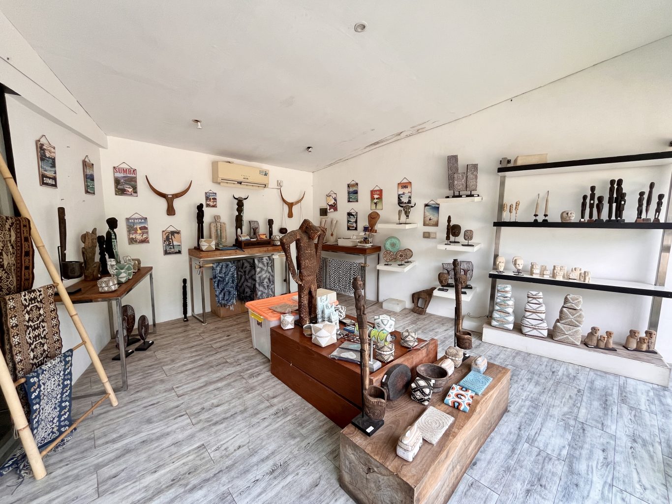 A bright, white-walled art shop interior in Sumba displays numerous traditional wooden carvings, woven textiles, and decorative artifacts on shelves and tables.