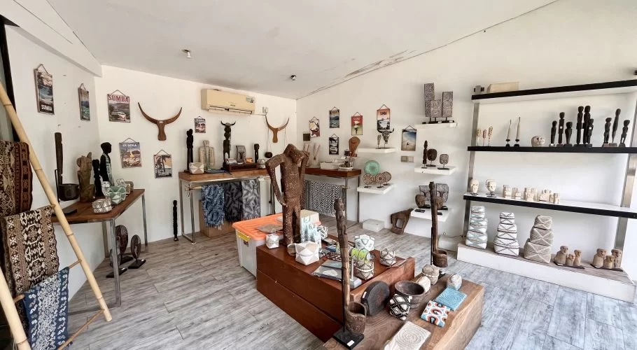 A bright, white-walled art shop interior in Sumba displays numerous traditional wooden carvings, woven textiles, and decorative artifacts on shelves and tables.