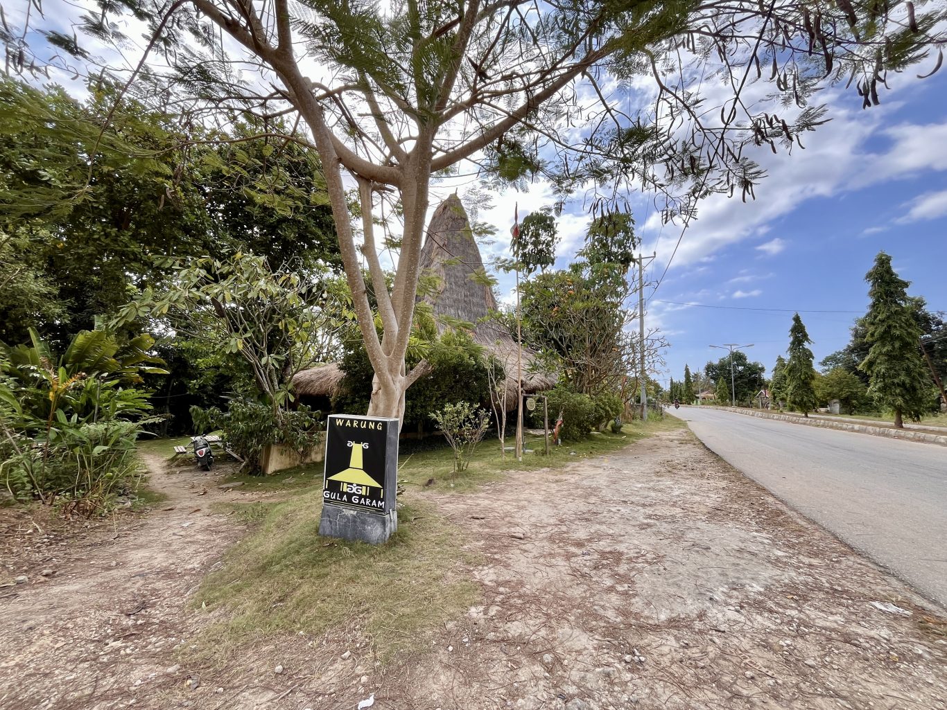 A traditional house-shaped restaurant with a tall thatched roof is nestled among green trees beside a paved road under a bright blue sky.