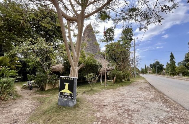 A traditional house-shaped restaurant with a tall thatched roof is nestled among green trees beside a paved road under a bright blue sky.