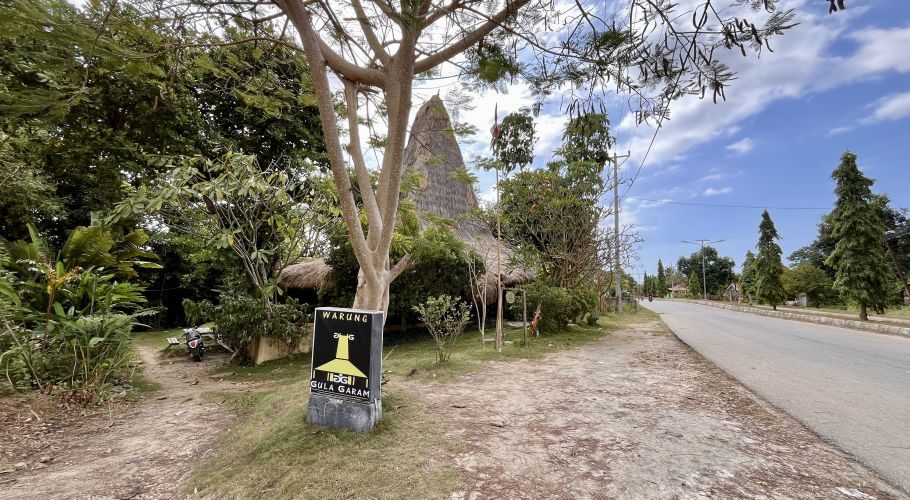 A traditional house-shaped restaurant with a tall thatched roof is nestled among green trees beside a paved road under a bright blue sky.